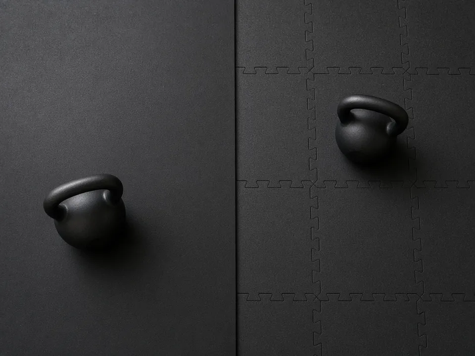 A plain black rubber mat beside black interlocking tiles, with a kettlebell resting on each.
